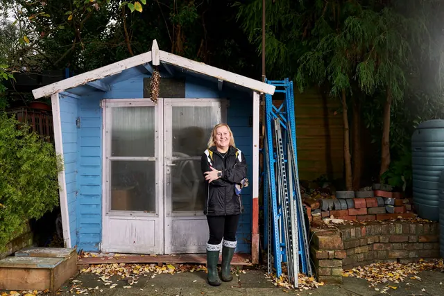 A happy woman stands in front of a blue shed, surrounded by garden equipment.